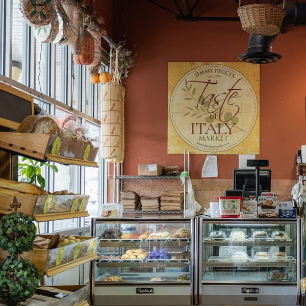 Interior of Taste of Italy Market with hanging cured meats, fresh bread, and pastry display cases.