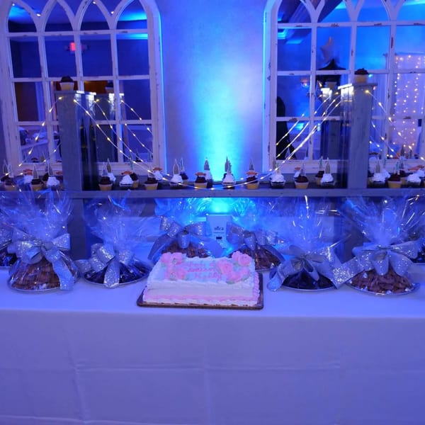 A dessert table with cupcakes, wrapped treats, and a pink and white cake under blue lighting.