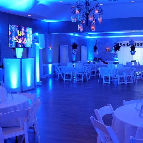 A banquet hall with blue lighting, round tables, white chairs, a large screen, and a photo booth.
