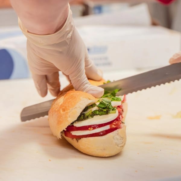 Gloved hands slicing a fresh Italian sandwich filled with mozzarella, tomatoes, and basil.