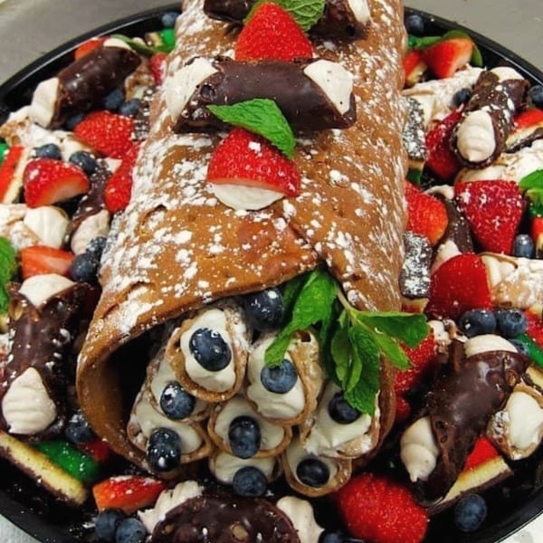 A large cannoli cake roll filled with mini cannolis, blueberries, strawberries, and mint leaves.