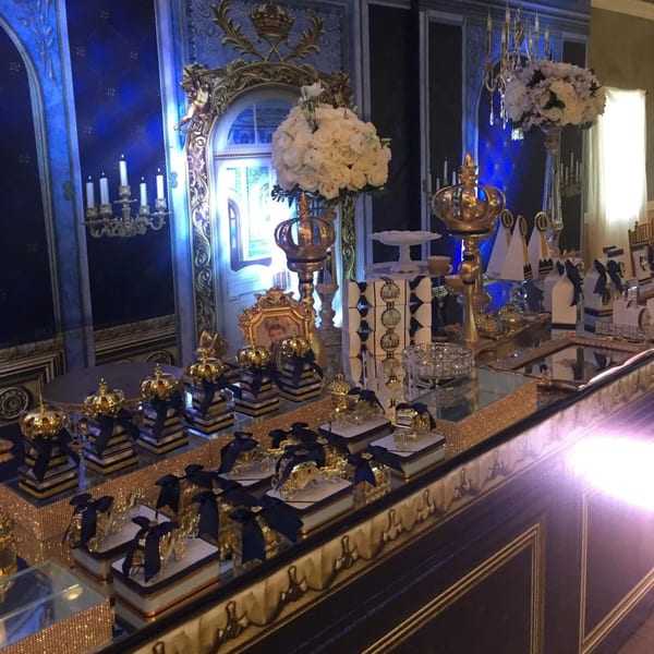 A luxurious blue and gold dessert table with white floral arrangements and crown decorations.