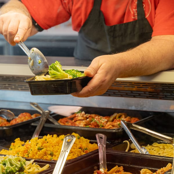 A chef serving Italian pasta and broccoli into a takeout container from a buffet.