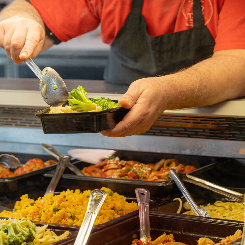 A chef serving Italian pasta and broccoli into a takeout container from a buffet.