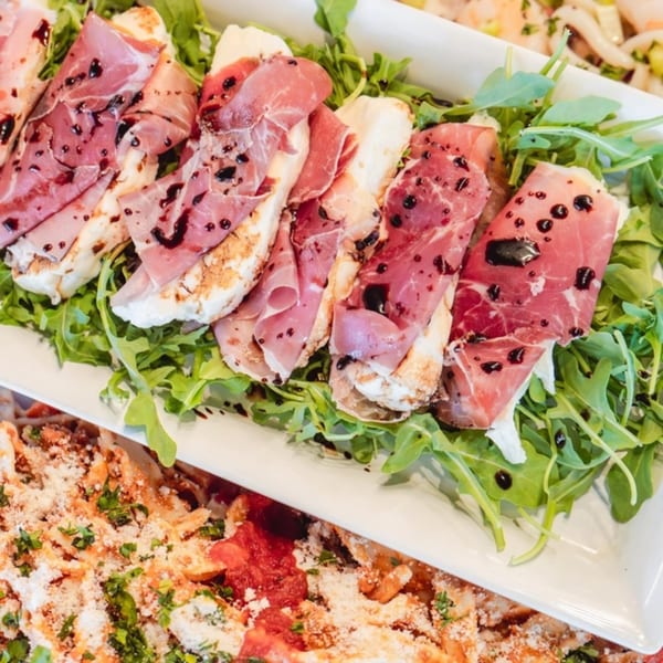 Italian bruschetta with prosciutto, mozzarella, arugula, and balsamic glaze, next to pasta.