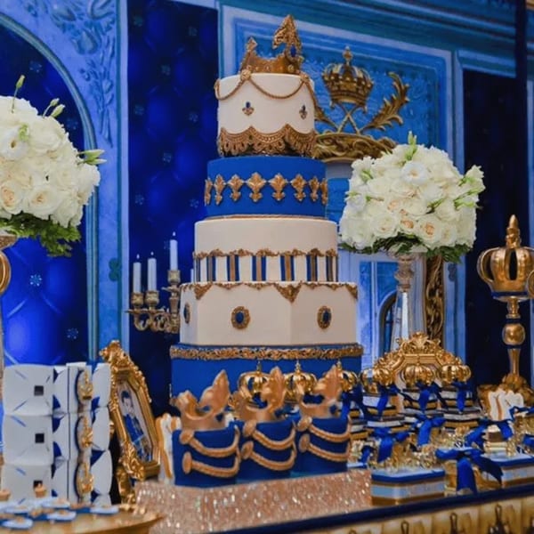 A lavish blue and gold tiered cake with crowns, surrounded by white floral arrangements and ornate decorations.