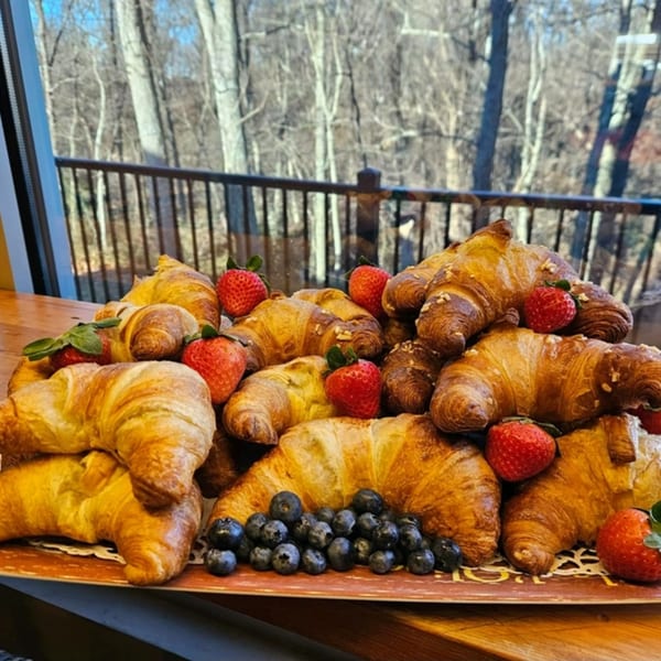 A platter of golden croissants, fresh strawberries, and blueberries on a table with a forest view.