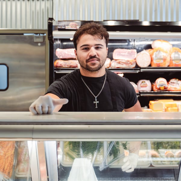 A deli worker in a black shirt and cross necklace stands behind a counter with meats and cheeses.