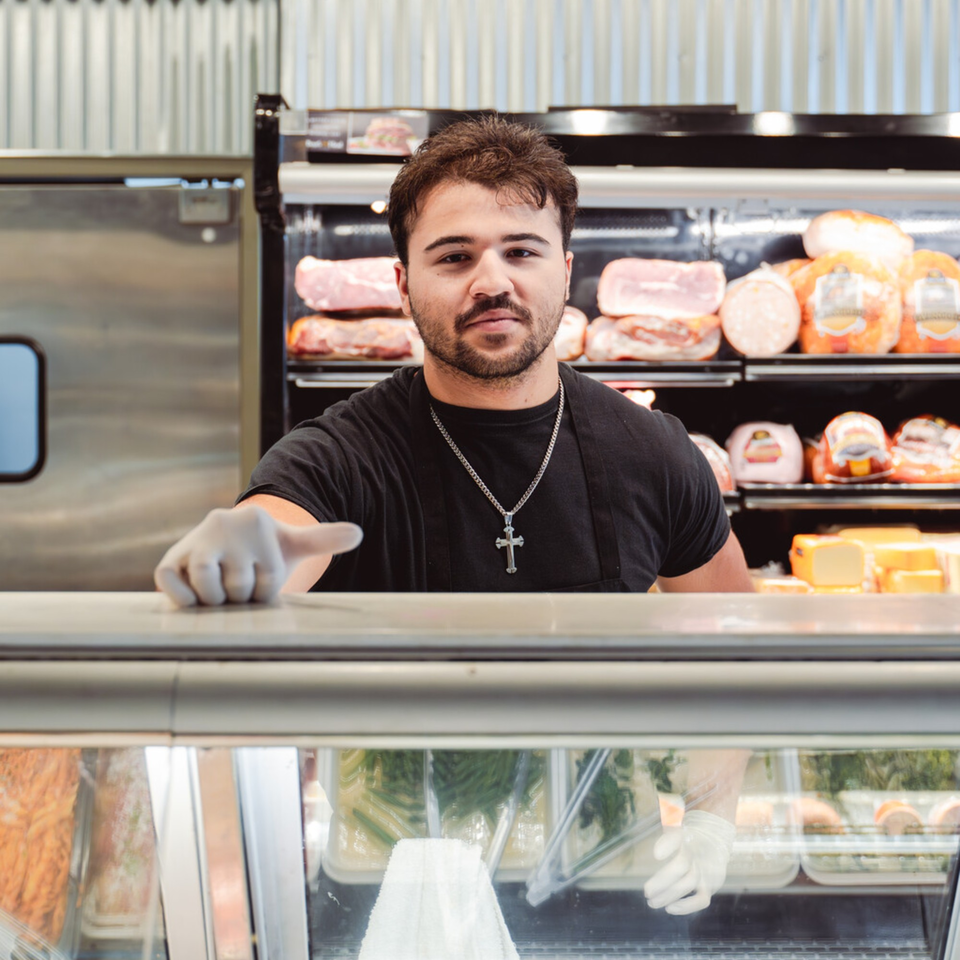 A deli worker in a black shirt and cross necklace stands behind a counter with meats and cheeses.