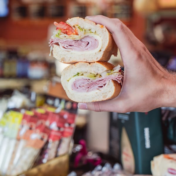 A hand holds a halved Italian sub sandwich filled with meats, cheese, lettuce, and tomato.